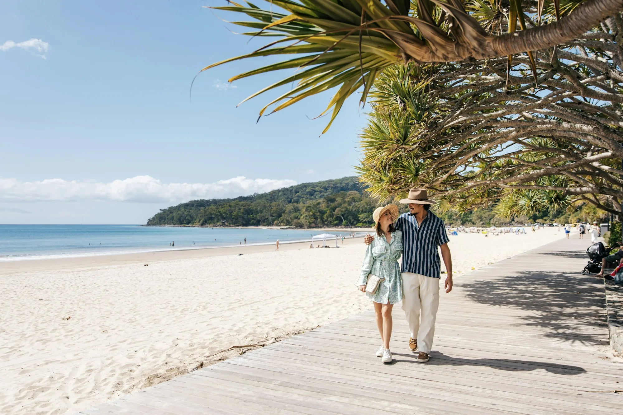 A couple in sun hats walking arm in arm along a timber beach boardwalk under pandanus trees